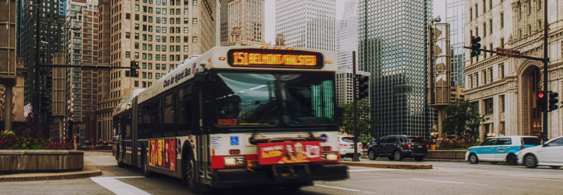 A city bus featuring an advertisement for personal injury attorneys on its side is driving through an urban area with tall buildings. The bus's display sign is lit, indicating its route. Several cars are on the road, and there are traffic lights and street signs visible.