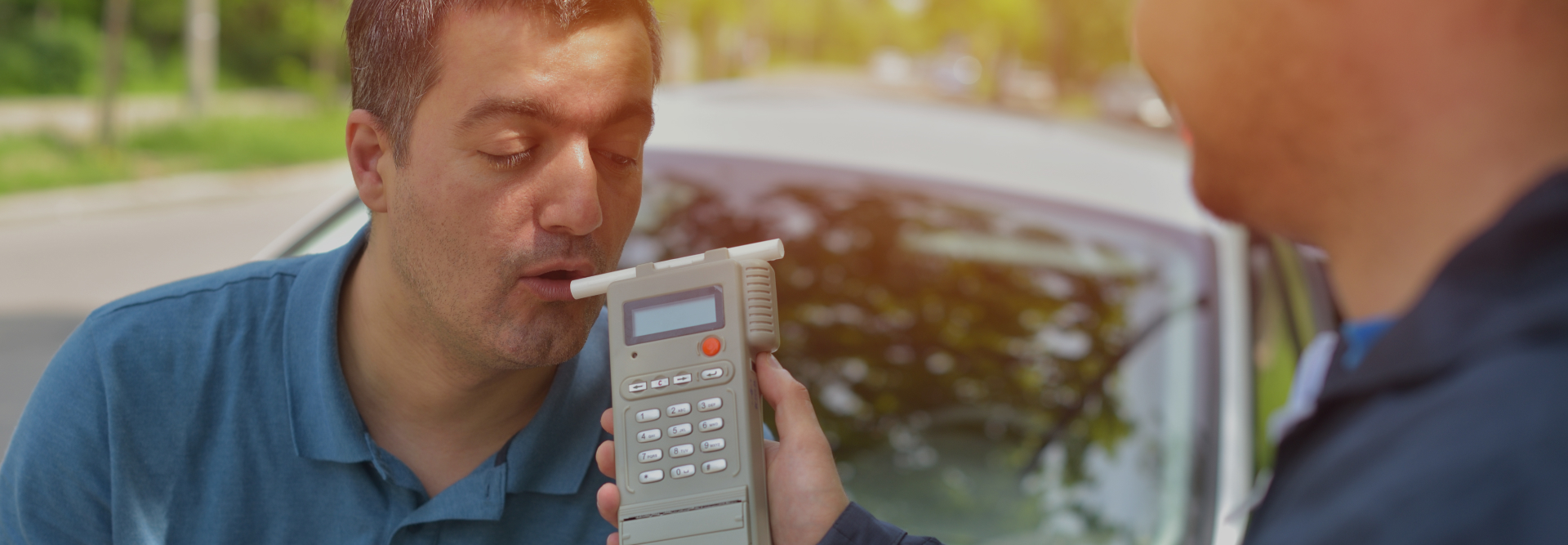 A man blows into a breathalyzer held by another person, possibly a police officer, outside near a car during the daytime. Trees and a blurred background indicate an outdoor setting. The scene suggests a roadside sobriety test, potentially involving vehicle accident attorneys.