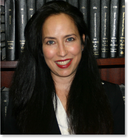 A woman with long, dark hair and red lipstick is smiling. She is wearing a dark blazer and white blouse, seated in front of a bookshelf filled with legal books often referenced by personal injury attorneys.