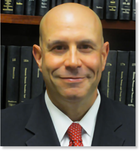A bald man wearing a dark suit, white shirt, and red patterned tie is smiling. He is posing in front of a bookshelf filled with black hardcover books, embodying the professionalism typical of injury lawyers.