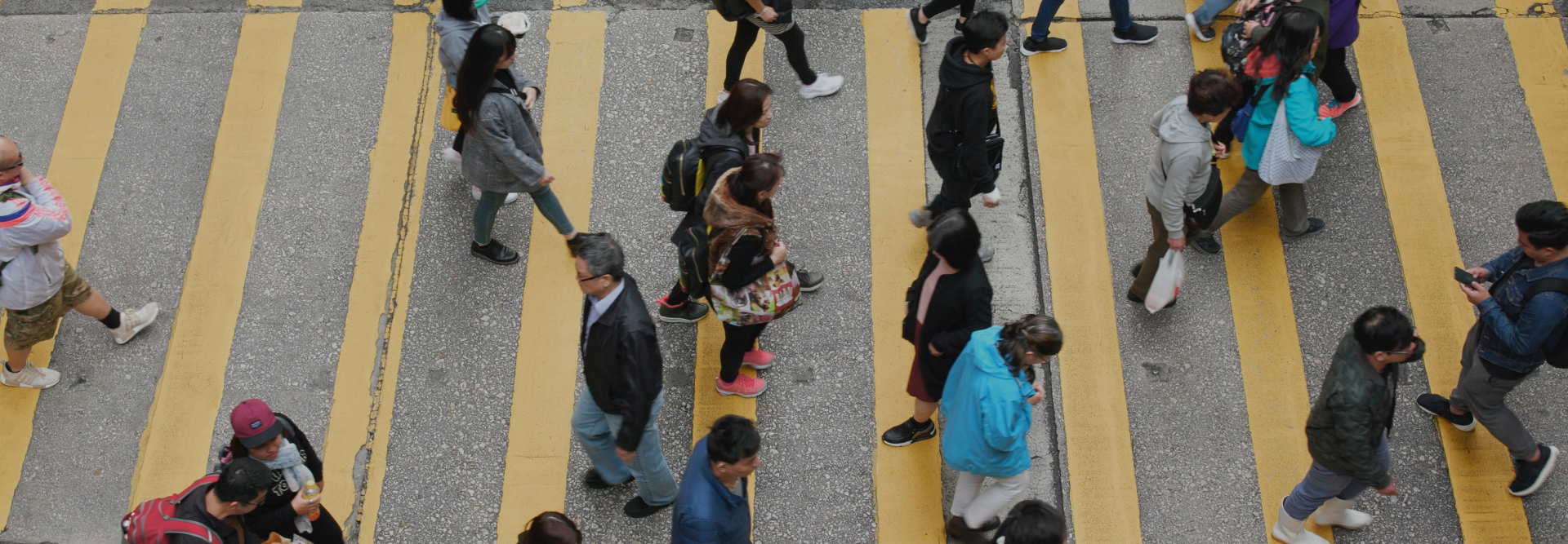 A crowd of people walking on a wide street with several yellow pedestrian crossing lines. They are dressed in various casual outfits, carrying bags or backpacks, and some are holding phones. The street is grey and appears to be in an urban area, where personal injury attorneys might often be needed.