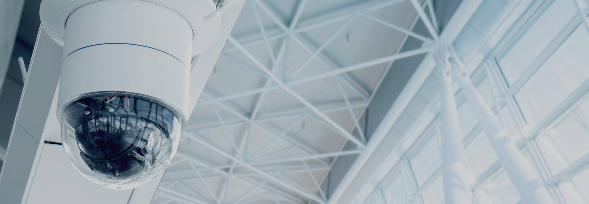 A close-up view of a ceiling-mounted security camera with a dome casing inside a modern building featuring large windows and a high, grid-patterned ceiling. The camera diligently observes the interior space, ensuring safety for everyone, including clients visiting their vehicle accident attorneys.