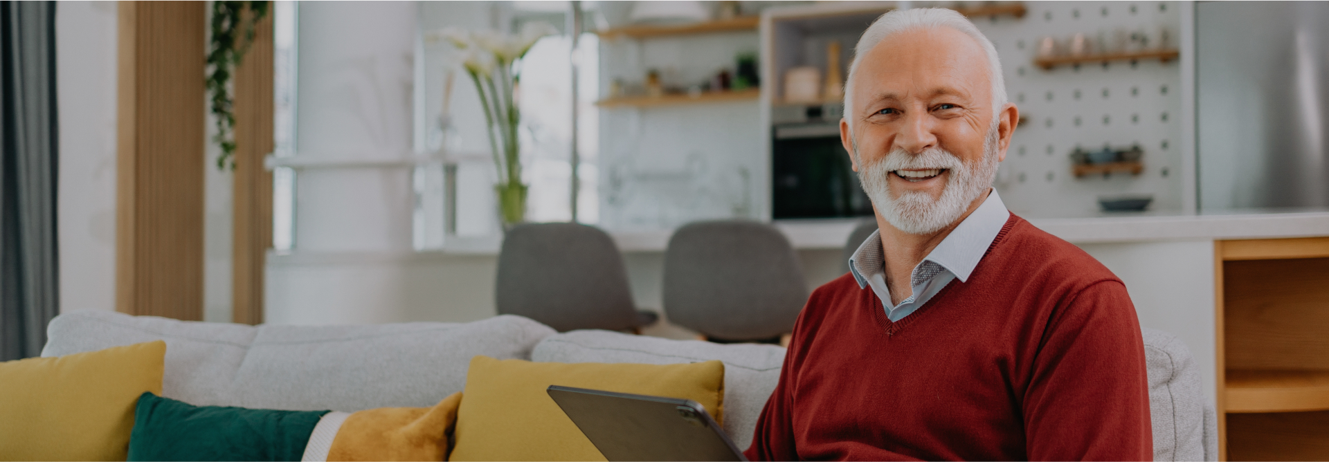 A smiling older man with white hair and a beard sits on a couch, holding a tablet and possibly reading about personal injury attorneys. He is wearing a red sweater over a collared shirt. The background features a modern living space with shelves, chairs, and various home decor elements.