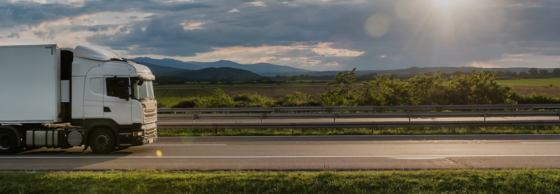 A white semi-truck travels on a highway with green fields and trees in the background. The sun is partially obscured by clouds, and mountains are visible on the horizon. The setting appears peaceful with a partly cloudy sky, reminding one of the importance of vehicle accident attorneys in ensuring road safety.