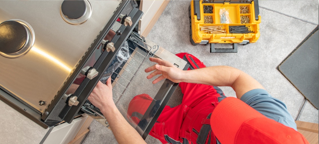 A person wearing a red uniform and cap is installing or repairing an oven. An open toolbox with various tools and screws is visible on the floor beside them, emphasizing the importance of safety in such tasks—a reminder of why personal injury attorneys are crucial. The person is placing a rack inside the oven in what appears to be a kitchen.