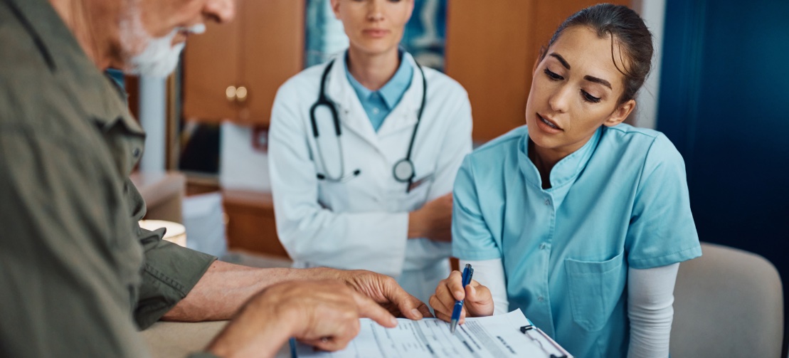 A nurse in blue scrubs is assisting an elderly man with filling out a medical form at a reception desk, while a female doctor in a white coat looks on. Nearby, a brochure for personal injury attorneys is prominently displayed in the medical office environment.