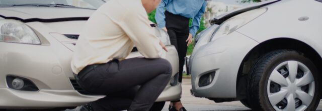 Two people inspect damage to their cars after a collision. One person is crouching down examining the front bumper of a silver car, while the other stands nearby, looking at the damage on their own vehicle. Both vehicles have visible damage, and they might soon need advice from vehicle accident attorneys.